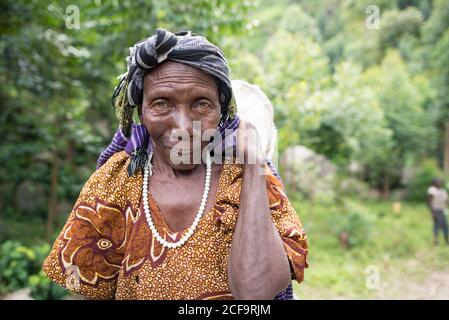 Ouganda - novembre, 26 2016: Femme africaine senior de corps entier regardant la caméra et le sac de transport sur le dos tout en marchant sur le chemin dans la forêt Banque D'Images