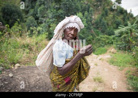 Ouganda - novembre, 26 2016: Femme africaine senior de corps entier regardant la caméra et le sac de transport sur le dos tout en marchant sur le chemin dans la forêt Banque D'Images