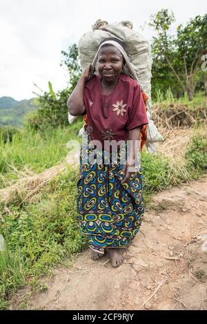 Ouganda - novembre, 26 2016: Femme africaine senior de corps entier regardant la caméra et le sac de transport sur le dos tout en marchant sur le chemin dans la forêt Banque D'Images