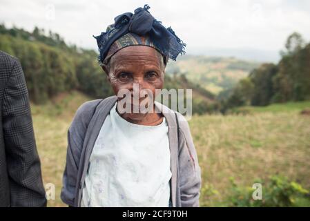 Ouganda - 26 novembre 2016 : femme africaine âgée et pensive vêtue de vêtements décontractés, regardant l'appareil photo avec un regard gentil sur des paysages flous étonnants de collines boisées vertes en journée Banque D'Images