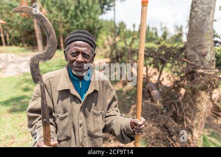 Ouganda - novembre 26 2016 : vieil homme africain avec hache et bâton regardant la caméra au milieu d'une forêt dans la campagne Banque D'Images