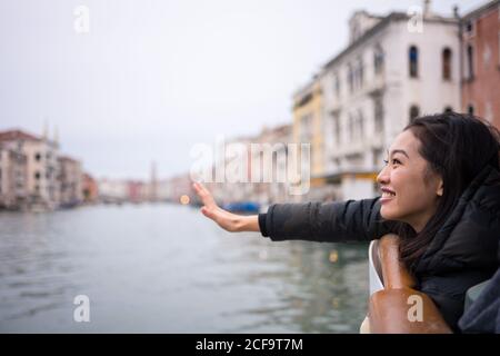 Vue latérale d'une dame asiatique de repos très joyeuse, vêlée de vêtements chauds souriant et en sortant du bateau sur la voie navigable au milieu de vieux bâtiments Banque D'Images