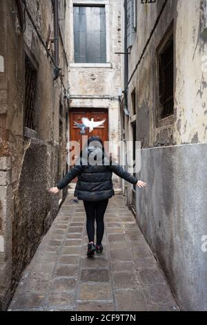 Vue arrière d'une femme sans visage à cheveux noirs vêque de vêtements chauds marche avec les bras levés sur une étroite ruelle rockée Banque D'Images