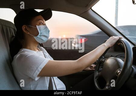 Vue latérale de la jeune femme asiatique dans des vêtements décontractés et masque de protection pour la prévention des coronavirus conduisant la voiture Banque D'Images