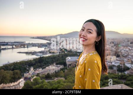 Vue latérale d'une femme asiatique optimiste souriant et regardant l'appareil-photo tout en visitant Castillo Gibraltar sur un fond flou de la ville côtière et le ciel ensoleillé à Malaga, Espagne Banque D'Images