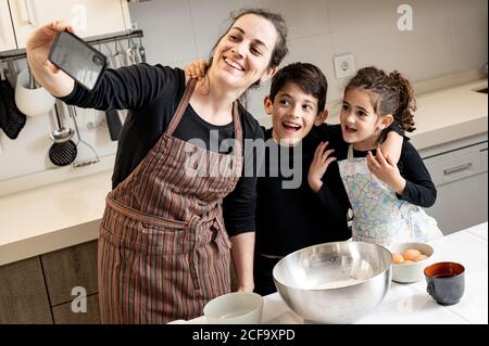 Une femme heureuse en tablier souriant et prenant le selfie avec un téléphone portable avec des enfants heureux tout en cuisinant des pâtisseries ensemble dans la cuisine confortable à la maison Banque D'Images