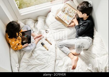 De dessus de petite fille regardant le dessin animé sur tablette et garçon lisant un livre intéressant pendant le week-end dans la chambre Banque D'Images