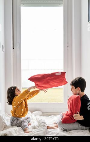 Vue latérale d'un frère et d'une sœur sympathiques assis sur confortable couverture près de la fenêtre et jouer avec des oreillers doux tout en ayant amusement pendant le week-end Banque D'Images