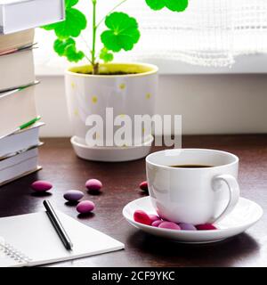 Une tasse de café blanche avec des bonbons multicolores se tient sur une table en bois, à côté d'une pile de livres et un carnet contre Banque D'Images
