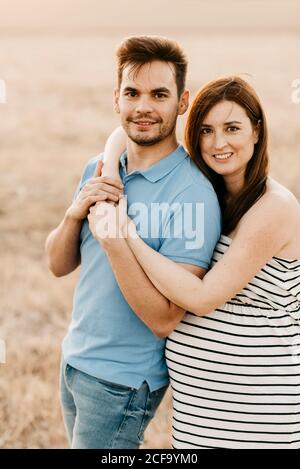 Bonne femme enceinte embrassant doucement homme smiley tout en regardant l'appareil photo dans un champ sec pendant le coucher du soleil dans la campagne Banque D'Images