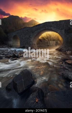 Paysage incroyable avec l'ancien pont en pierre traversant une rivière peu profonde contre montagnes majestueuses et ciel coloré au coucher du soleil en automne soir campagne Banque D'Images