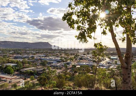 Vue sur la ville d'Alice Springs depuis la colline d'Anzac. Montagnes MacDonnell chaîne en arrière-plan. Soleil brillant parmi les feuilles de l'arbre à l'heure du coucher du soleil. Nuageux s Banque D'Images