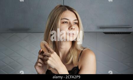 Jeune belle femme lissant ses cheveux blonds à la maison - arrière-plan gris flou Banque D'Images