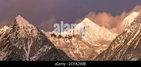 Pics de montagne pointus partiellement couverts de neige et entourés de brouillard sous un ciel pittoresque et nuageux rose et violet à la tombée de la nuit en hiver Banque D'Images