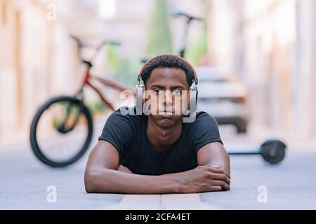 Jeune homme afro-américain concentré portant un casque d'écoute pour écouter de la musique en étant allongé sur une route asphaltée dans la rue Banque D'Images