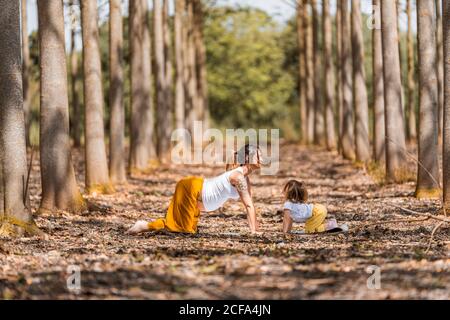 Vue latérale de la femme enceinte adulte et de la petite fille en chemises blanches et pantalons jaunes regardant les uns les autres et en riant tout en étirant le corps sur le sol pendant l'exercice du yoga dans la glade parmi les arbres dans la forêt pendant la journée ensoleillée Banque D'Images