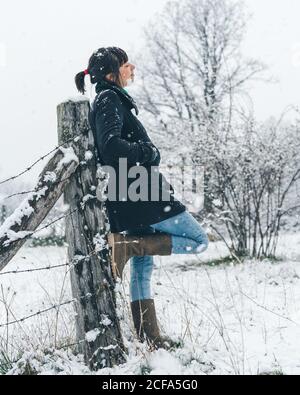 Vue latérale d'une femme décontractée et décontractée debout sur le terrain se pencher vers l'arrière et le pied sur une clôture en bois avec un fil barbelé et profiter d'une journée de neige tranquille Banque D'Images