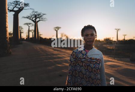 Madagascar - 6 JUILLET 2019: Femme ethnique confiante en tenue exotique multicolore debout en pente par de grands baobabs en rayons du soleil au crépuscule Banque D'Images