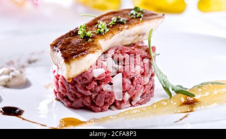 Gros plan de steak tartare juteux avec oignon blanc servi avec morceau de foie gras glacé sur plaque blanche avec sauces Banque D'Images