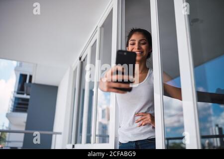 Belle brunette femme hispanique prenant selfie sur téléphone mobile et se tenant entre les fenêtres de balcon Banque D'Images