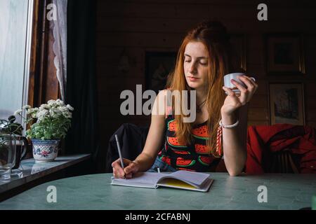 Femme adulte pensant assis à une table dans une pièce confortable et écrivant dans un carnet Banque D'Images