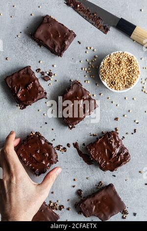 Vue de dessus de la personne coupée non reconnaissable mains tenant maison savoureux biscuits cuits coupés en carrés et recouverts de chocolat noir glaçage et graines de quinoa sur parchemin Banque D'Images