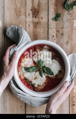 Vue de dessus de la personne anonyme mettant Hot Bowl plein de Délicieux poulet aux feuilles de parmesan et de basilic rôties à la tomate sauce sur la table de bois près des épices et des ustensiles Banque D'Images