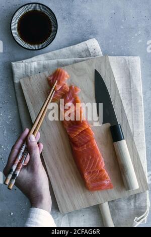 De la récolte ci-dessus personne anonyme tenant un morceau de saumon avec baguettes et trempette dans la sauce à la table servie Banque D'Images