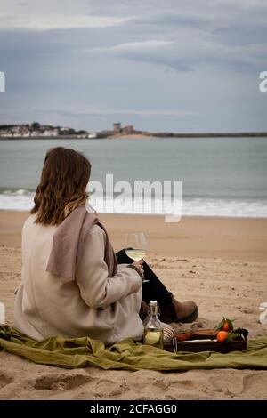 Vue arrière d'une femme méconnue sous un manteau élégant couverture près de la bouteille, un verre de vin blanc et un plateau avec tarte aux mandarines et pain au fromage et saucisses en vous reposant sur la plage le week-end Banque D'Images