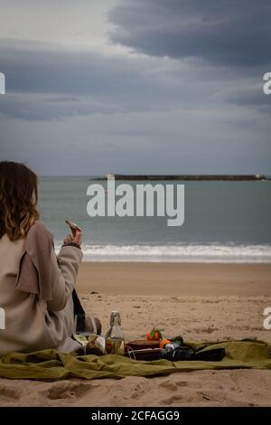 Vue arrière d'une femme méconnue sous un manteau élégant couverture près de la bouteille, un verre de vin blanc et un plateau avec tarte aux mandarines et pain au fromage et saucisses en vous reposant sur la plage le week-end Banque D'Images