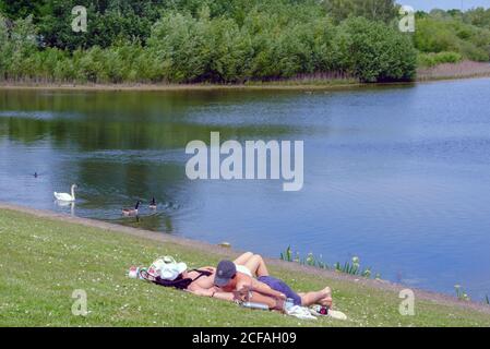 Un jeune couple se trouve sur l'herbe à côté de l'eau au réservoir du Lido de Ruislip avec des chaussures et des vestes. Cygnes et oies nageant dans le lac. Nord-Ouest de Londres. Banque D'Images