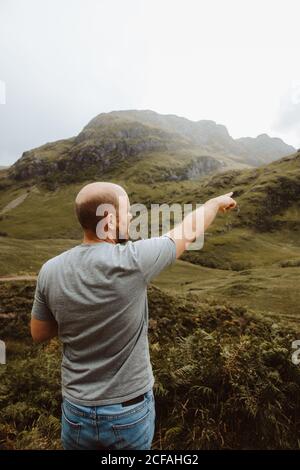 Vue arrière de mâle chauve se tenant dans la vallée verte Contemplant la vue pittoresque et montrant avec l'index à Glencoe Banque D'Images