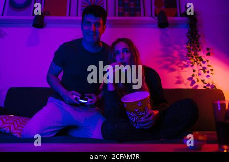Petit ami souriant jouant à la console de jeux vidéo tout en étant assis près d'excités petite amie mangeant du pop-corn sur un canapé confortable dans l'appartement au néon léger Banque D'Images