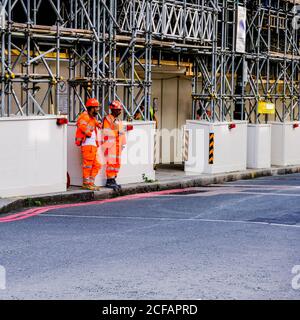 Deux constructeurs de combinaisons orange et de chapeaux durs debout sous Échafaudage temporaire à Londres, Royaume-Uni Banque D'Images