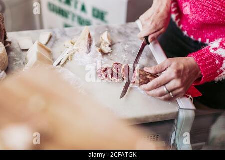 De dessus anonyme personne coupant de délicieux saucisses sur le comptoir près fromage tout en travaillant dans un magasin d'alimentation local Banque D'Images