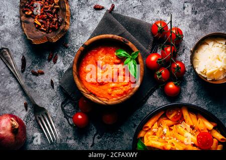 D'en haut de la sauce arrabiata colorée dans un bol en bois décoré avec basilic frais près des tomates cerises fraîches et des bols avec du fromage et tomates séchées et pâtes fraîches délicieuses décorées avec tomates cerises mûres sur une table de sorbets avec une serviette grise Banque D'Images