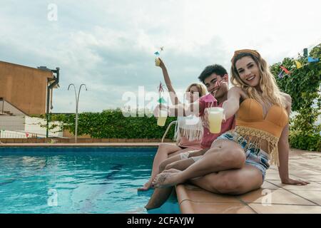 Groupe d'amis qui ont une fête au bord de la piscine pendant qu'ils dansent, rient et boivent des cocktails Banque D'Images