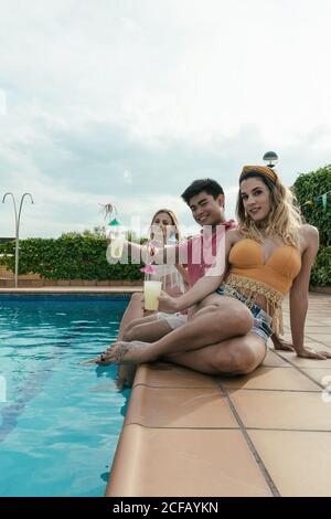 Groupe d'amis qui ont une fête au bord de la piscine pendant qu'ils dansent, rient et boivent des cocktails Banque D'Images