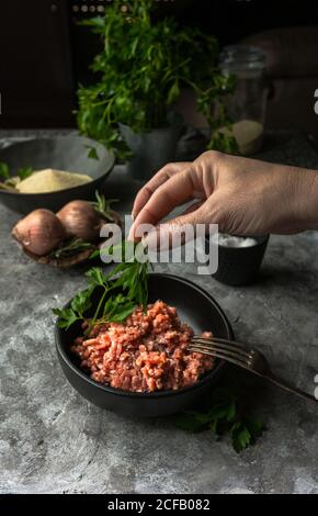 personne méconnaissable mettant du persil frais dans un bol avec du cru haché viande pendant la préparation du déjeuner Banque D'Images
