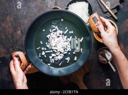 Vue de dessus de Crop anonyme femme de ménage secouer la casserole et le mélange oignon tout en cuisant des nouilles avec du poulet pour le dîner Banque D'Images