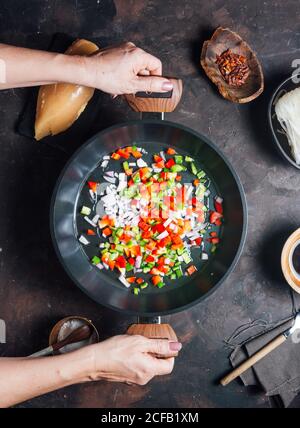 Vue de dessus de Crop anonyme femme de ménage secouer la casserole et le mélange poivron et oignon hachés pendant la cuisson des nouilles avec du poulet pour dîner Banque D'Images