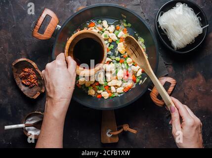 De dessus de la récolte anonyme femme versant la sauce de soja dans la casserole avec le poulet haché et les légumes tout en préparant de délicieuses nouilles dans la cuisine Banque D'Images