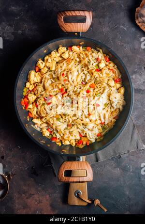 Vue de dessus des nouilles sautées épicées appétissantes avec poulet viande et légumes sur une poêle placée sur rustique foncé tableau Banque D'Images