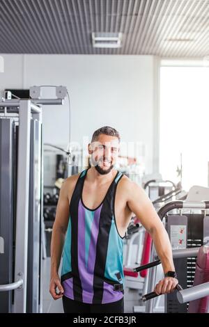 Homme barbu musclé dans les vêtements de sport regardant l'appareil photo en position debout bras croisés et inclinés à la lumière sur les équipements sportifs salle de sport contemporaine Banque D'Images