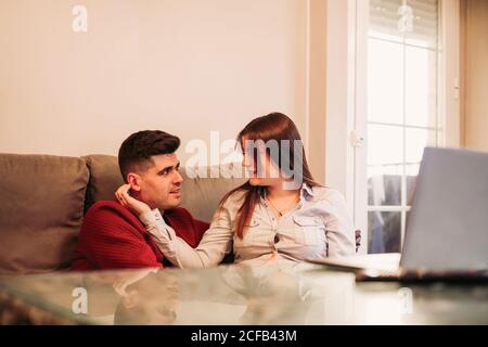 Jeune homme en rouge cavalier assis avec femme enceinte dedans blouse beige sur le canapé et toucher la bosse de bébé tout en dépensant ensemble sur l'ordinateur hiver à la maison Banque D'Images