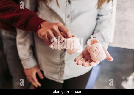 Crop jeune femme enceinte dans des vêtements décontractés debout à côté du mari et ensemble tenant des chaussons roses bébé avec des étoiles Banque D'Images