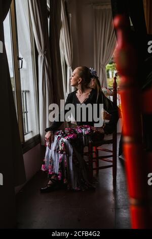 Vue latérale d'une jolie danseuse pensive en jupe fleurie pour le flamenco assis et donnant sur la fenêtre Banque D'Images