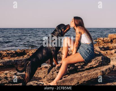 Vue latérale d'une femme tatouée et branchée assise sur la pierre et embrassant le chien Rottweiler tout en passant du temps sur le rivage rocheux Banque D'Images