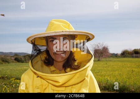 Gardien positif en costume de protection jaune et masque souriant et regarder la caméra en se tenant dans le champ vert apiary dans la journée ensoleillée d'été Banque D'Images