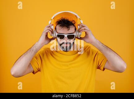 Un homme barbu dans une tenue élégante écoutant de la musique sur un casque blanc contemporain sur fond jaune Banque D'Images